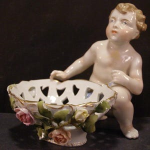 May include: A white porcelain bowl with a cherub figurine holding the bowl. The bowl is decorated with gold trim and has a delicate openwork design. The bowl is also decorated with three pink and white roses.