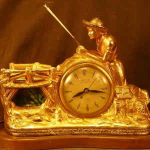 May include: A gold-colored vintage clock featuring a fisherman with a fishing rod, sitting atop a detailed landscape. The clock face displays the time, with the brand name "United" visible.