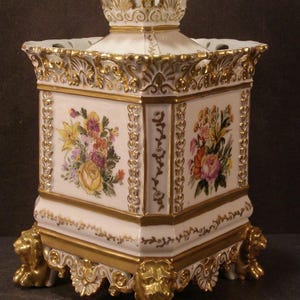 May include: A square porcelain vessel featuring white surfaces with hand-painted floral bouquets in yellow, pink, and green. The piece is adorned with ornate gold-gilded trim, decorative relief borders, and four sculpted lion paw feet at the base.