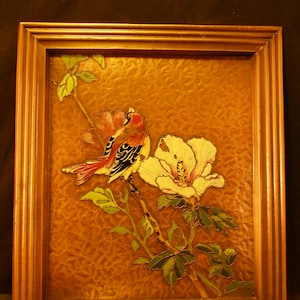 May include: A framed artwork featuring a colorful bird perched on a branch with a white flower. The background is a textured copper color.