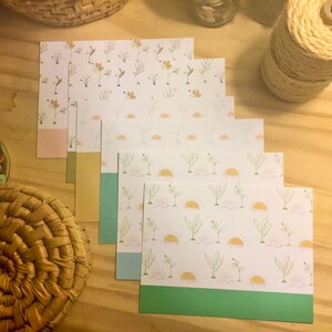 May include: A stack of stationery cards with floral and sun designs. The cards feature a white background with green, orange, and yellow illustrations. Each card has a solid-colored bottom border in shades of pink, green, blue, and yellow. A wicker basket and twine spool are in the background.
