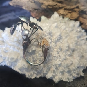 May include: A silver and gold ring with a delicate design resembling a flower with a pearl in the center. The ring is set on a white crystal background.