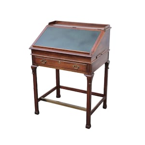 May include: A brown wooden slant-top desk featuring a dark green leather writing surface, a single drawer with two brass pulls, and four legs connected by a horizontal brass footrest bar. The desk has a hinged lid and a keyhole.
