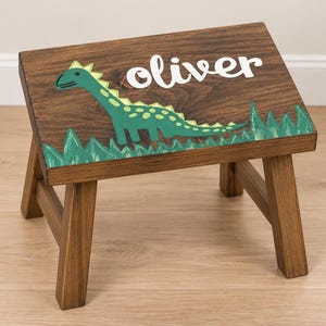 May include: A dark brown wooden step stool featuring a painted green dinosaur graphic and the name oliver in white script lettering. The stool has four sturdy legs and a rectangular top surface with visible wood grain patterns throughout the piece.