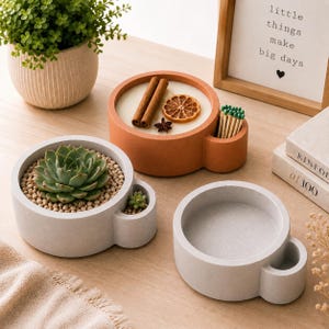 May include: Three round, cup-shaped containers made of smooth material. One light gray container holds a green succulent plant in small pebbles. One terracotta-colored container holds a candle with cinnamon sticks, a dried orange slice, and matches.