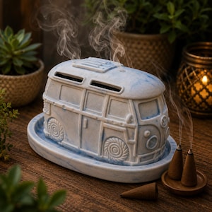 May include: A light blue and white marbled ceramic incense burner shaped like a vintage van. The van sits on a matching oval tray and emits thin wisps of smoke from its roof vents. Three brown cone-shaped incense pieces are placed nearby.