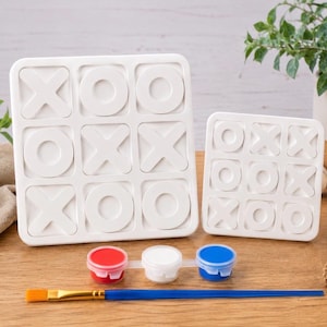 May include: Two white square tic-tac-toe game boards with raised X and O symbols. The set includes a strip of three small paint pots in red, white, and blue, along with a single paintbrush featuring a blue handle and a yellow bristle tip.