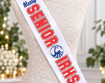 May include: A white graduation sash featuring the name Alexis in blue script, the word SENIOR in red block letters, an eagle logo in blue, the letters IRHS in red, and the text class of 2027 in blue with a graduation cap icon.