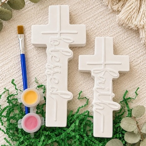 May include: Two white ceramic crosses featuring embossed script text reading blessed and faith. The set includes a blue-handled paintbrush and two small circular paint pots containing yellow and pink paint, arranged for a craft project.