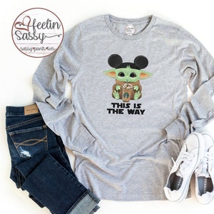 May include: Grey long-sleeved shirt featuring a green character wearing Mickey Mouse ears and holding a coffee cup. The text "This is the way" is printed below the character.