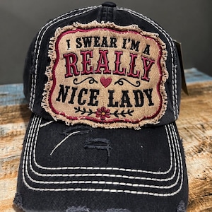 May include: A black baseball cap with a distressed finish and white stitching. A rectangular tan patch with frayed edges is centered on the front, featuring the embroidered text "I SWEAR I'M A REALLY NICE LADY" in black and dark pink with a small heart and flower.