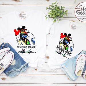 Puede incluir: Dos camisetas blancas con un dinosaurio de colores que lleva orejas de Mickey Mouse y sostiene un globo rojo. El dinosaurio está de pie sobre un cartel que dice "Wrong Park".