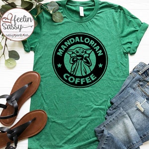 Puede incluir: Camiseta verde con un gráfico en blanco y negro de un personaje de la serie de televisión Mandalorian tomando café. El gráfico tiene la forma de un logotipo de una cafetería con el texto "Mandalorian Coffee".