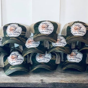Könnte beinhalten: Ein Stapel von Camouflage-Trucker-Mützen mit einem weißen Patch, auf dem "Sun Sip Sand Siesta" in orangefarbenen und braunen Buchstaben steht.