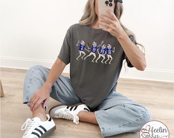 May include: Dark gray t-shirt featuring a skeleton football team in blue and white uniforms. The shirt has the numbers 4, 17, 73, and 10 on the jerseys. A white and blue "Buffalo" hat is also visible.