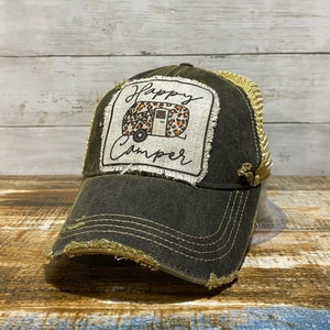 May include: A dark green distressed trucker hat with a gold mesh back. The front of the hat features a square patch with a camper van design and the text "Happy Camper" in white.