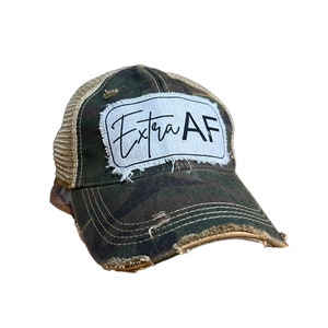 May include: A camouflage baseball cap with a tan mesh back and distressed detailing. The front displays a white patch with the words "Extra AF" in black script. The cap has a worn appearance with frayed edges.