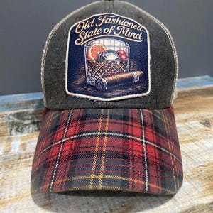 Pode incluir: Boné de caminhoneiro com coroa cinza escuro e aba xadrez vermelha e preta. O patch frontal apresenta uma ilustração de coquetel com o texto "Old Fashioned State of Mind."