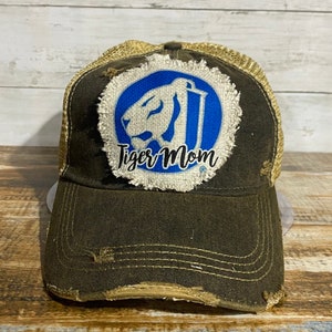 May include: Distressed brown trucker hat with a tan mesh back. The front features a blue and white patch with a tiger design and the words "Tiger Mom" in black script. The hat has a pre-worn, vintage look.