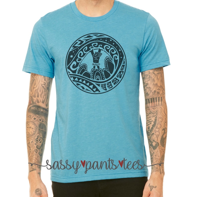 May include: A light blue t-shirt with a black and white graphic of a Polynesian-style design featuring a person standing on a wave. The design includes a circle with waves and stars around the outside. The text "Sassy Pants Tees" is printed on the bottom of the shirt.