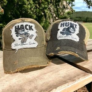 May include: Two distressed trucker hats with a cartoon character and the text "32nd Weapons Squadron" on the front. The hat on the left says "HACK" and the hat on the right says "HUNT".