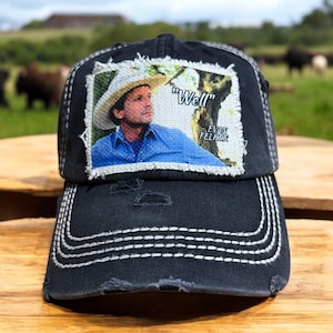May include: A black distressed baseball cap with a white patch featuring a man wearing a cowboy hat and the text "Well - Evan Felker".
