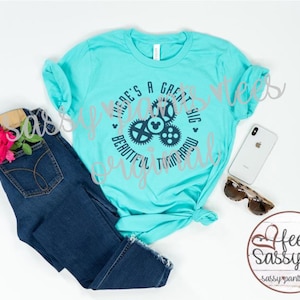 May include: A turquoise t-shirt with the text "THERE'S A GREAT BIG BEAUTIFUL TOMORROW" in a circular design with gears and a Mickey Mouse head. The shirt is paired with blue jeans, sunglasses, and a smartphone.