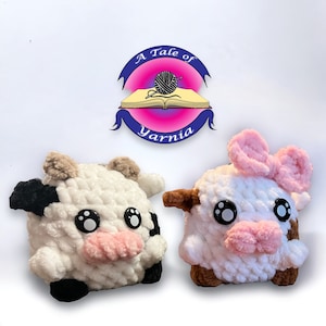 May include: Two handmade crochet cow plushies. One cow is white with black spots and a pink nose. The other cow is white with brown spots and a pink bow and nose. The logo "A Tale of Yarnia" is above the cows.