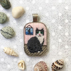 May include: A black and white cat with green eyes is embroidered on a pink fabric pendant. The pendant is framed in a silver metal frame and has a blue and white picture of a house on the back.