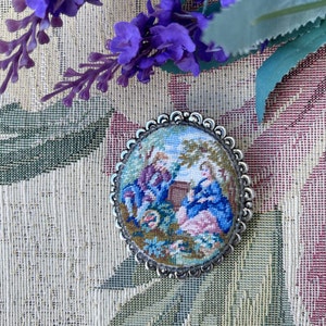 Könnte beinhalten: Eine ovale Brosche aus Silber mit einem detaillierten Blumendesign und einer Szene mit zwei Personen in einem Garten. Die Szene ist mit bunten Fäden gestickt, darunter Blau, Rosa und Grün.