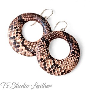 May include: A pair of hoop earrings with a snakeskin pattern in shades of brown, tan, and black. The earrings have a circular design with a silver-colored hook. The text "T's Studio Leather" is at the bottom.