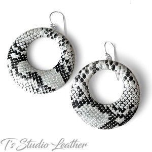 May include: A pair of large, round hoop earrings made of white and black snakeskin leather. The earrings have a silver hook closure.