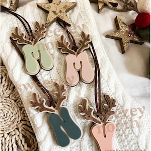 May include: Four wooden reindeer ornaments with the letter "M" in various pastel colors: mint green, pink, teal, and peach. Each ornament has brown antlers and a leather cord for hanging. The ornaments are on a white knitted surface, with gold star decorations and text that reads "Sweezy North".