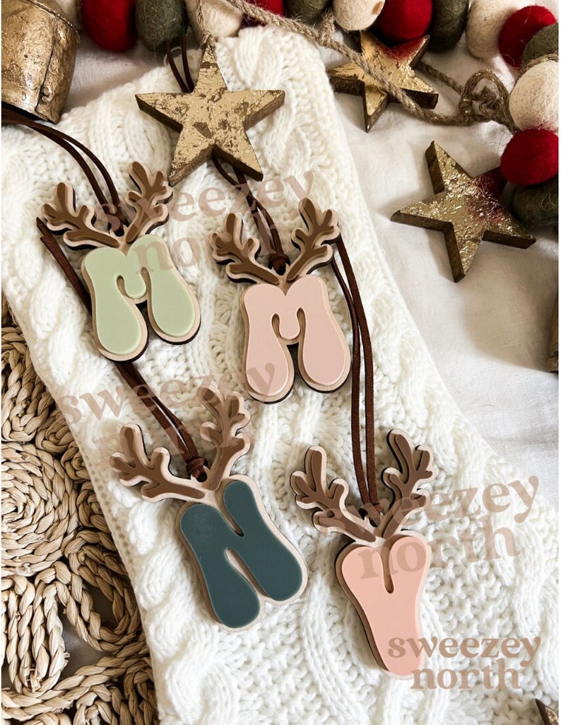 May include: Four wooden reindeer ornaments with the letter "M" in various pastel colors: mint green, pink, teal, and peach. Each ornament has brown antlers and a leather cord for hanging. The ornaments are on a white knitted surface, with gold star decorations and text that reads "Sweezy North".