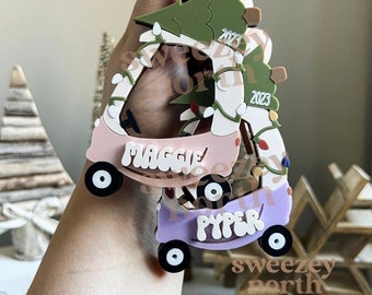 May include: A Christmas ornament shaped like a golf cart, in pink and purple, with the names "Maggie" and "Pyper" on it. The cart has a green Christmas tree on top, with the year "2023" and white string lights. The wheels are black.