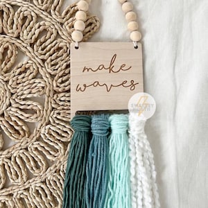 May include: Wooden wall hanging with the text "make waves" and four tassels in shades of green, blue, and white. The hanging is suspended by a string of wooden beads. The background is a neutral linen fabric.