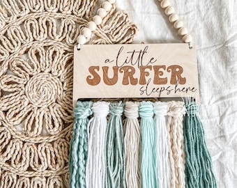 May include: Wooden sign with the text "a little surfer sleeps here" in brown lettering with a decorative fringe of teal, white, and beige yarn.