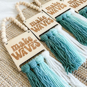 May include: Wooden signs with the text "make waves" in a retro font, hanging from wooden bead garlands. Each sign has a multi-colored tassel attached.