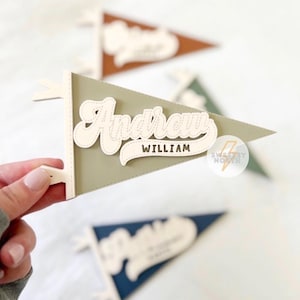 Könnte beinhalten: Ein personalisierter Wimpel in Salbeigrün mit dem Namen "Andrew William" in weißer Schrift. Weitere Wimpel in Braun, Grün und Blau sind im Hintergrund zu sehen. Die Wimpel sind ein dekorativer Artikel.