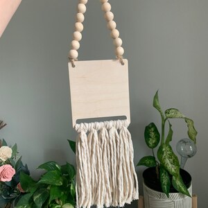 May include: A wooden sign with a natural wood finish and a white tassel fringe. The sign is hung with a string of wooden beads.
