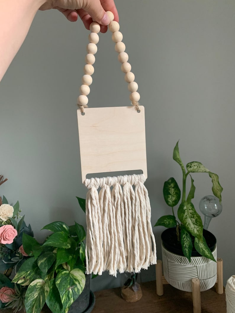 May include: A wooden sign with a natural wood finish and a white tassel fringe. The sign is hung with a string of wooden beads.