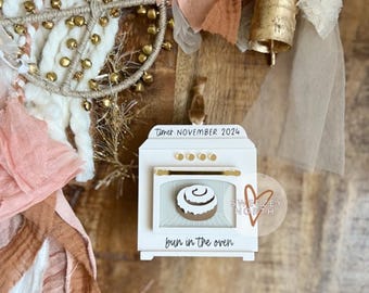May include: A white oven-shaped decoration with the text "Timer NOVEMBER 2024" and "bun in the oven." The oven has a gold handle and knobs, and a cinnamon roll is visible inside. The decoration is part of a larger arrangement.