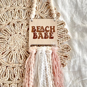 May include: Wooden sign with the text "Beach Babe" in a retro font, with a beaded garland and pink, white, and beige tassels hanging below.