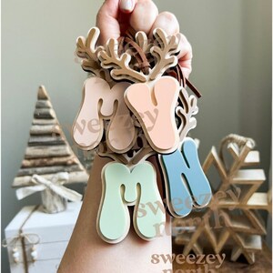 May include: A hand holds a collection of wooden ornaments. Each ornament features the letters "M", "A", and "M" in pastel colors, with a reindeer antler design. The text "sweezey north" is visible on the ornaments.