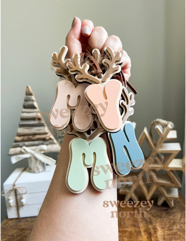 May include: A hand holds a collection of wooden ornaments. Each ornament features the letters "M", "A", and "M" in pastel colors, with a reindeer antler design. The text "sweezey north" is visible on the ornaments.
