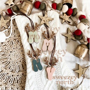 May include: Four wooden Christmas ornaments shaped like reindeer with the letters "M", "N", and "Y" in pastel colors. The ornaments hang from brown leather cords. The background includes a white knitted stocking, star-shaped ornaments, and a garland of red and green felt balls.