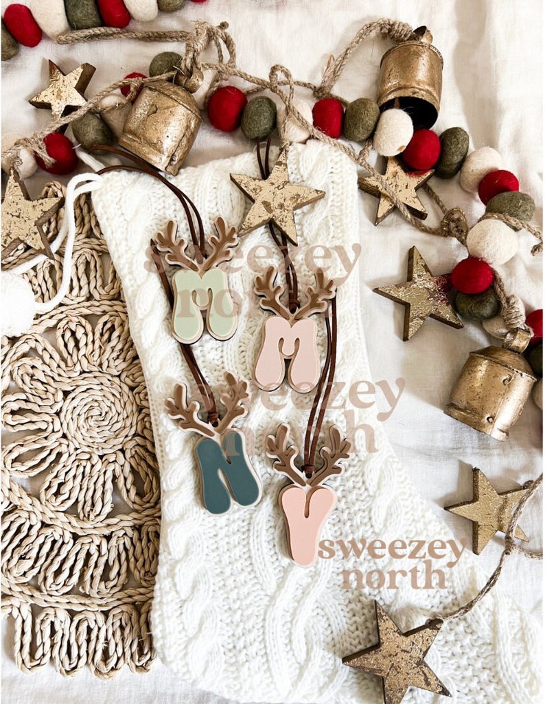 May include: Four wooden Christmas ornaments shaped like reindeer with the letters "M", "N", and "Y" in pastel colors. The ornaments hang from brown leather cords. The background includes a white knitted stocking, star-shaped ornaments, and a garland of red and green felt balls.