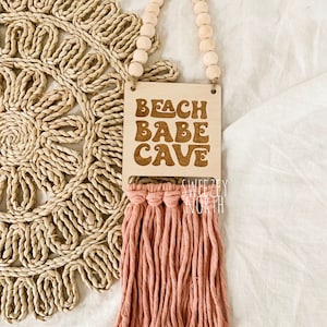 May include: Wooden sign with the text "Beach Babe Cave" in a retro font, hanging from a string of wooden beads. The sign has a pink tassel hanging from the bottom.