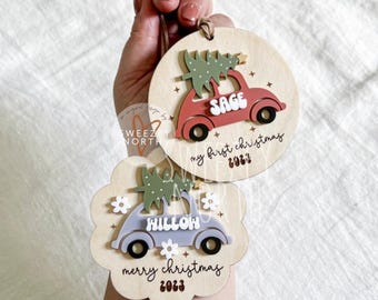 May include: Two wooden Christmas ornaments. One is round with a red car carrying a green tree, the name "Sage", and the text "my first christmas 2023". The other is a scalloped shape with a blue car, the name "Hilloh", and the text "merry christmas 2023".