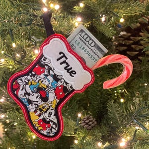 Può includere: Un ornamento a forma di calza natalizia con bordo rosso e anello nero per appenderla. La calza è riempita con una banconota da cento dollari e un bastoncino di zucchero. La calza presenta un colorato motivo di Topolino e la parola "True".